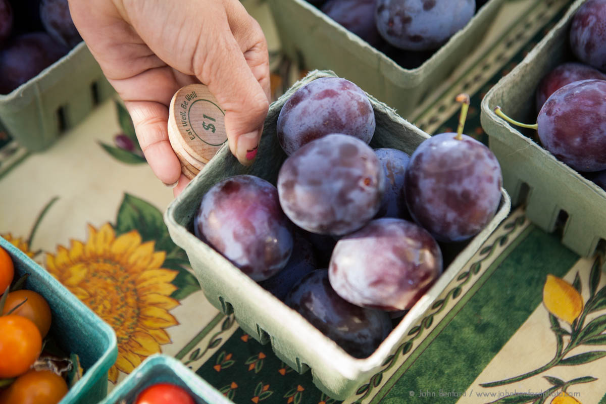 close up of hand holding pint of plums and wooden dollar coins at farmers market, credit: John Benford